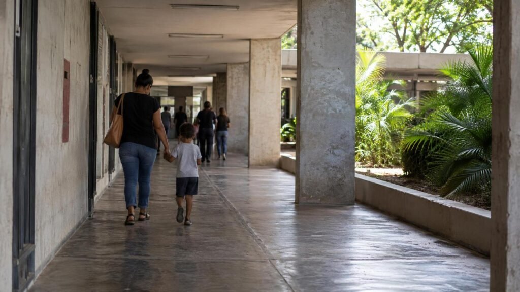 Responsável e criança caminham de mãos dadas por um corredor externo de serviço público, vistos por trás e sem rostos identificáveis.