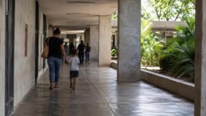 Responsável e criança caminham de mãos dadas por um corredor externo de serviço público, vistos por trás e sem rostos identificáveis.