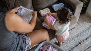Responsável e criança organizam roupas em uma sala doméstica vivida, vistos de cima e sem rostos identificáveis.